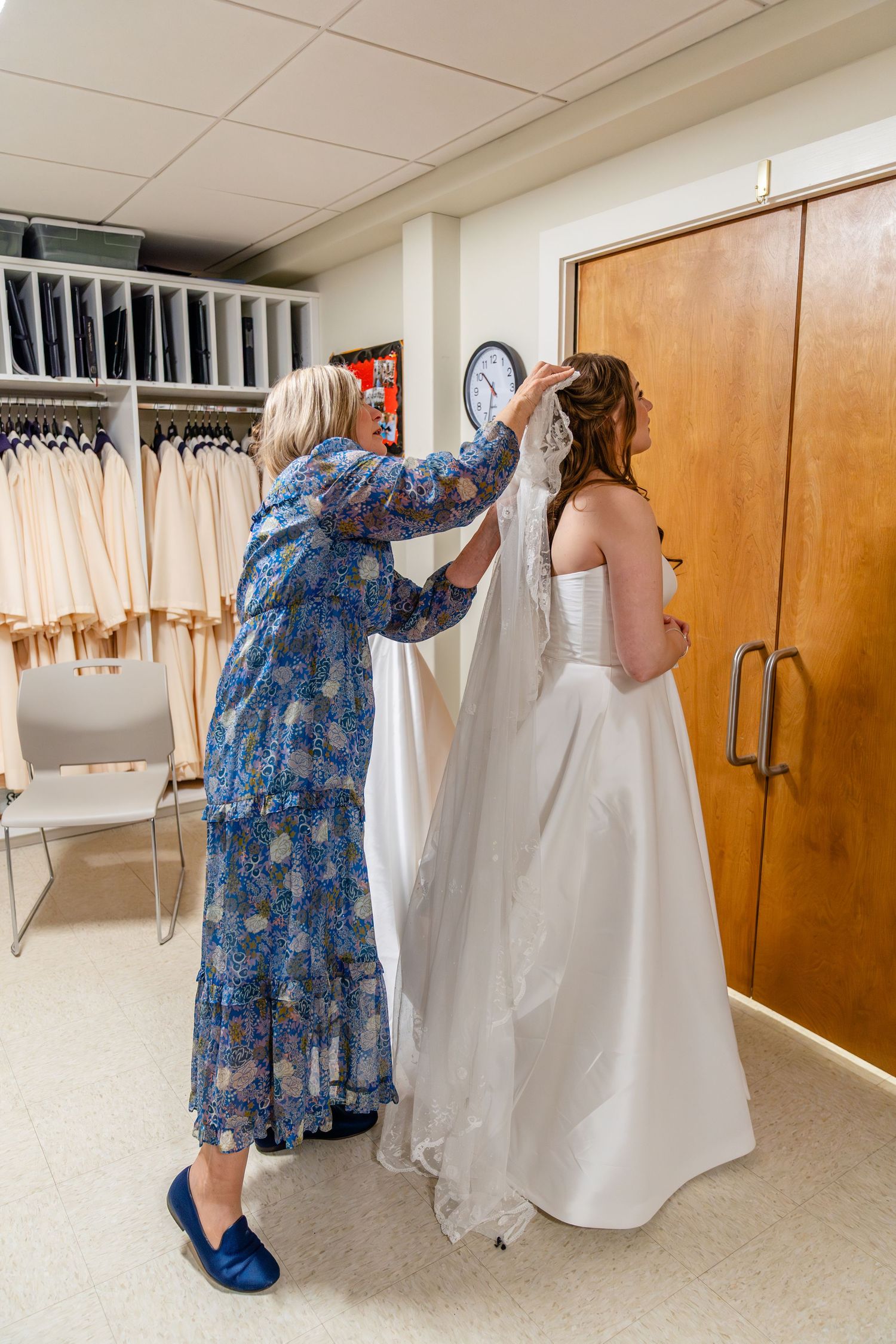 A woman in a floral blue dress helps adjust a white wedding gown in a dressing room with hanging robes visible.