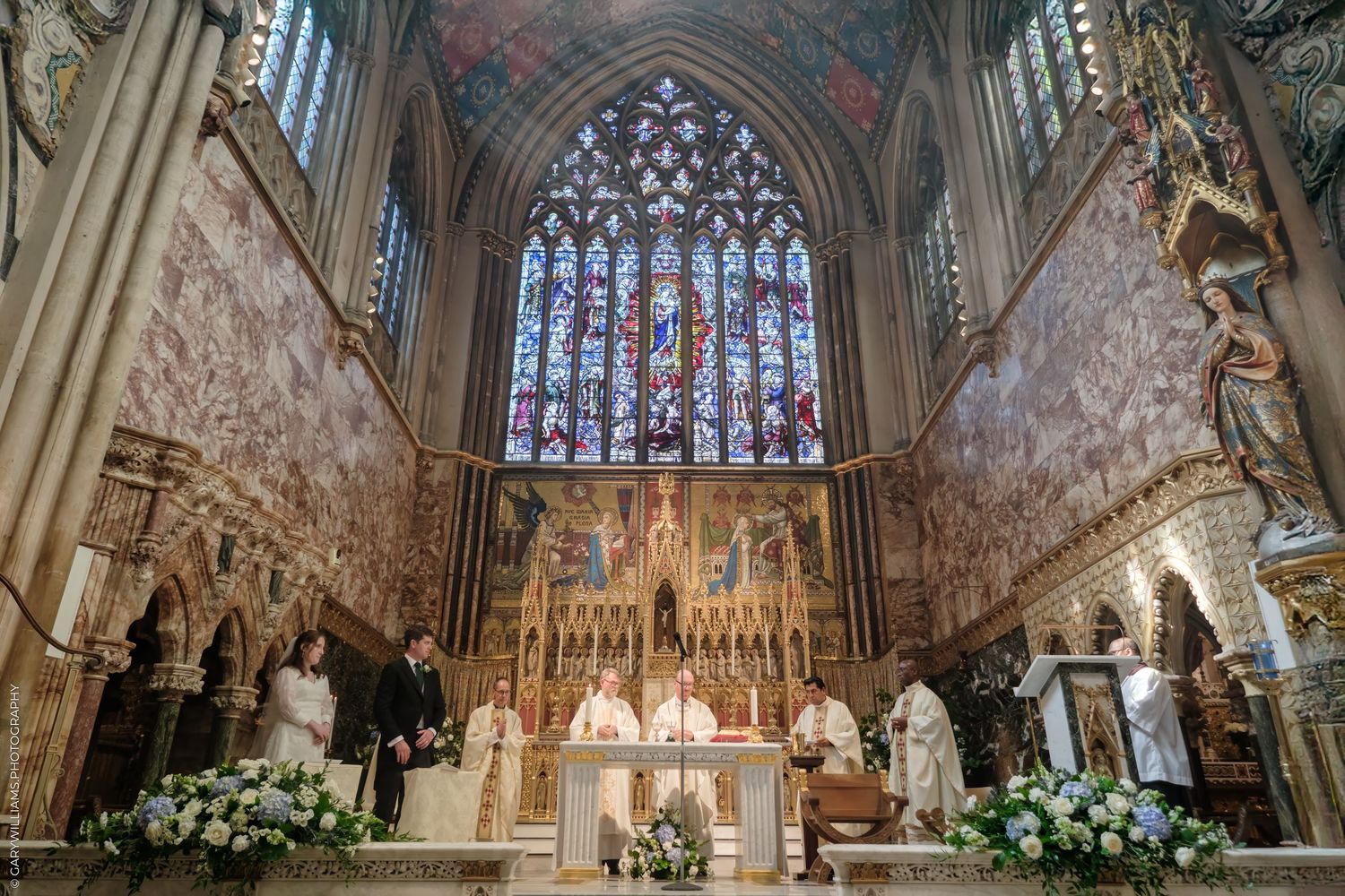 Gothic cathedral altar with ornate stonework, stained glass window, and white floral arrangements for a wedding ceremony.