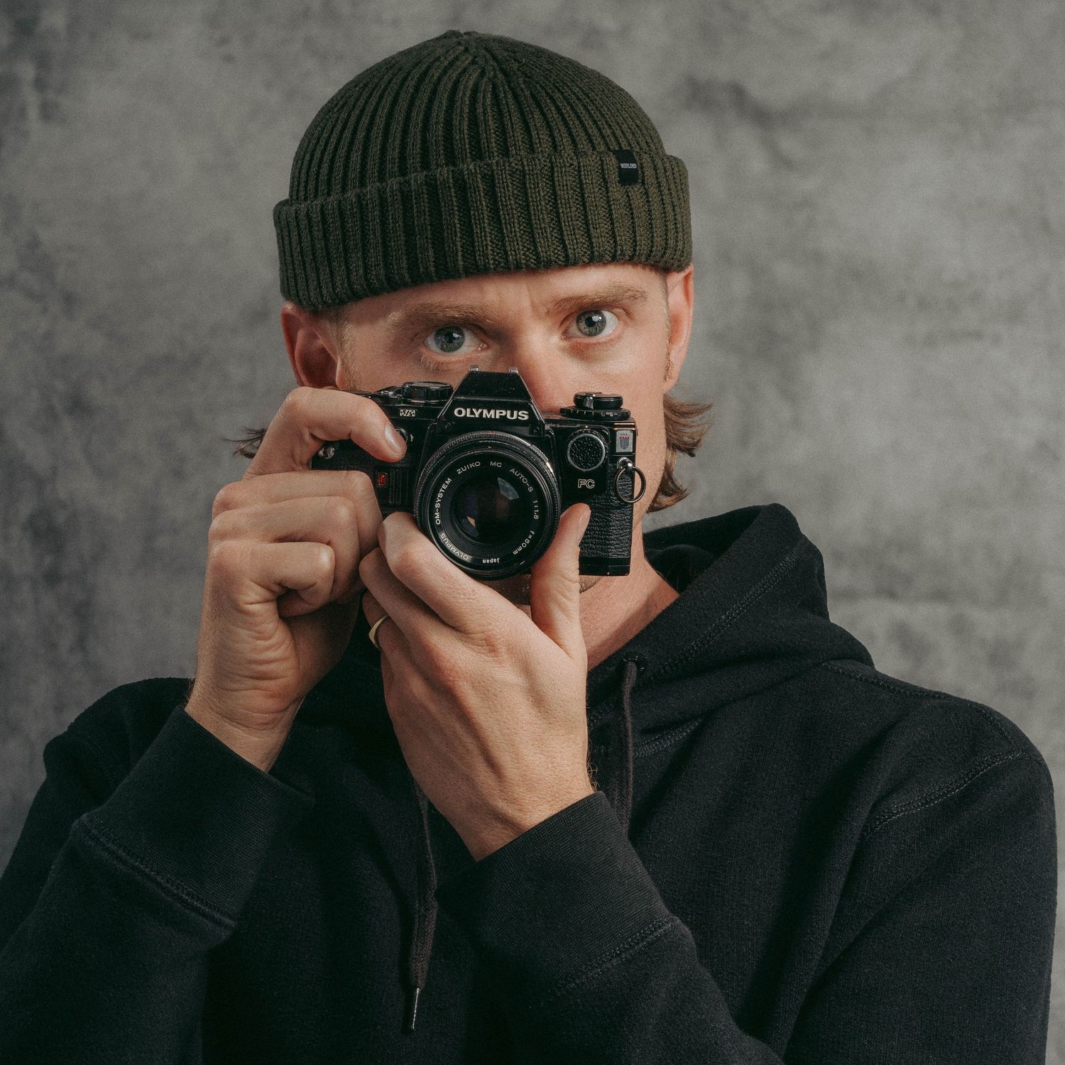 Photographer in dark beanie and hoodie taking photo with vintage camera against gray background.
