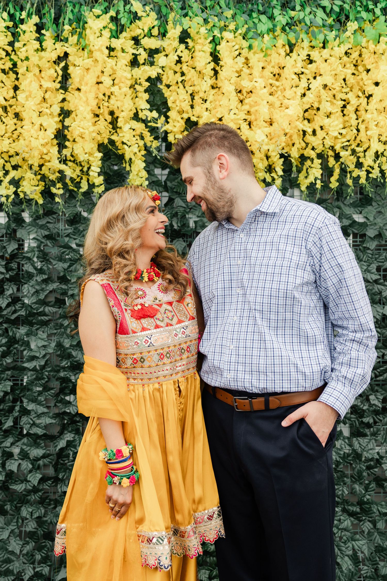 A couple shares loving looks in front of bright yellow flowers and green foliage during an engagement photo session.
