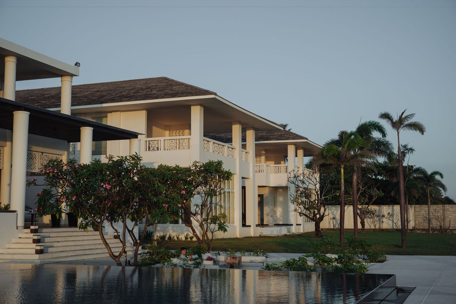Elegant white colonial villa with palm trees reflected in a tranquil infinity pool at dusk.
