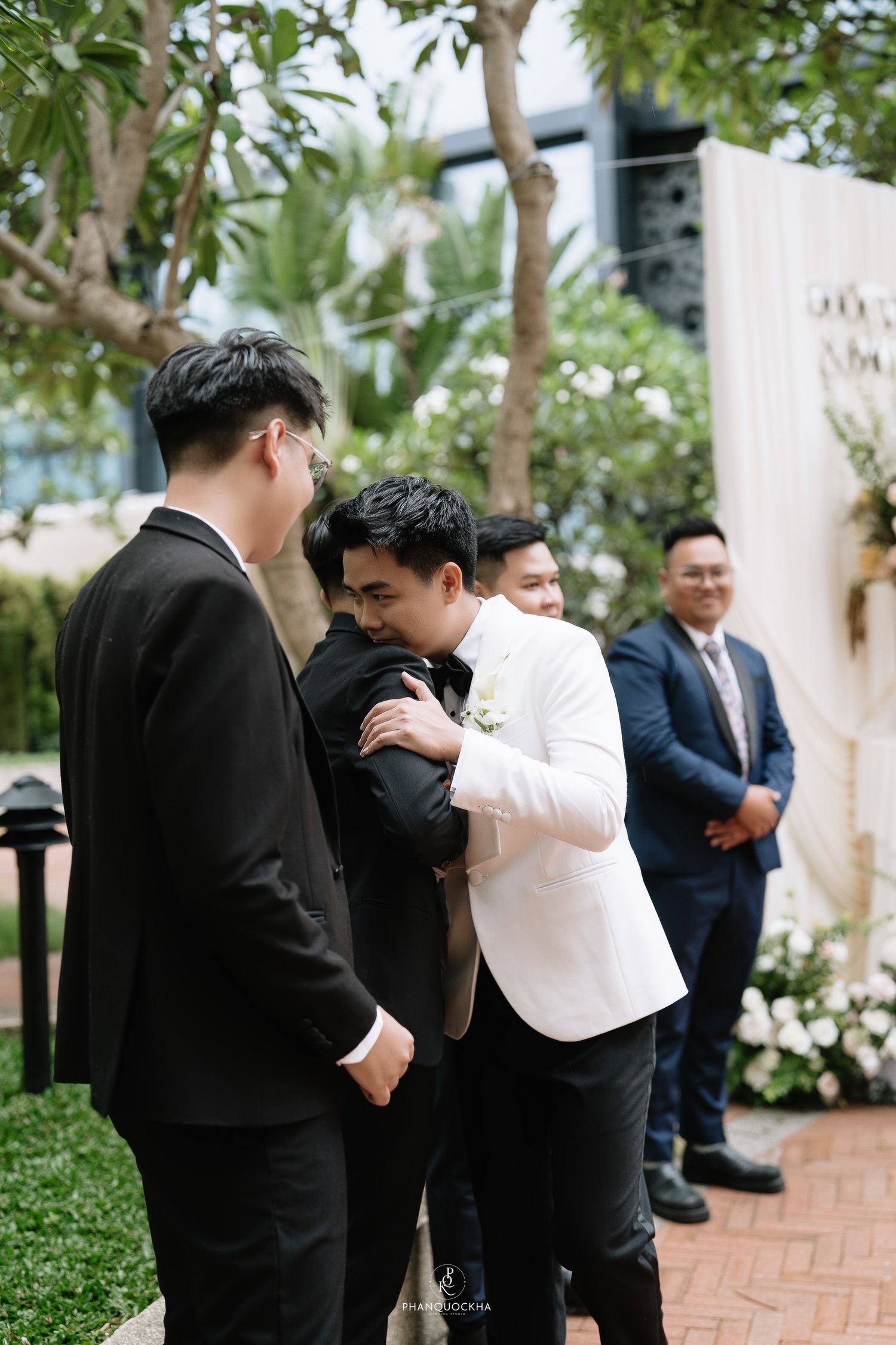 Groom in white tuxedo embracing a guest while groomsmen in black suits stand nearby at outdoor wedding.