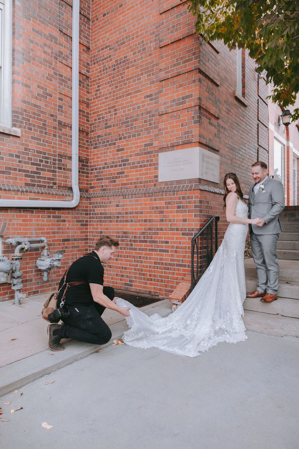 A photographer arranges a long lace wedding dress train against a brick wall building exterior.