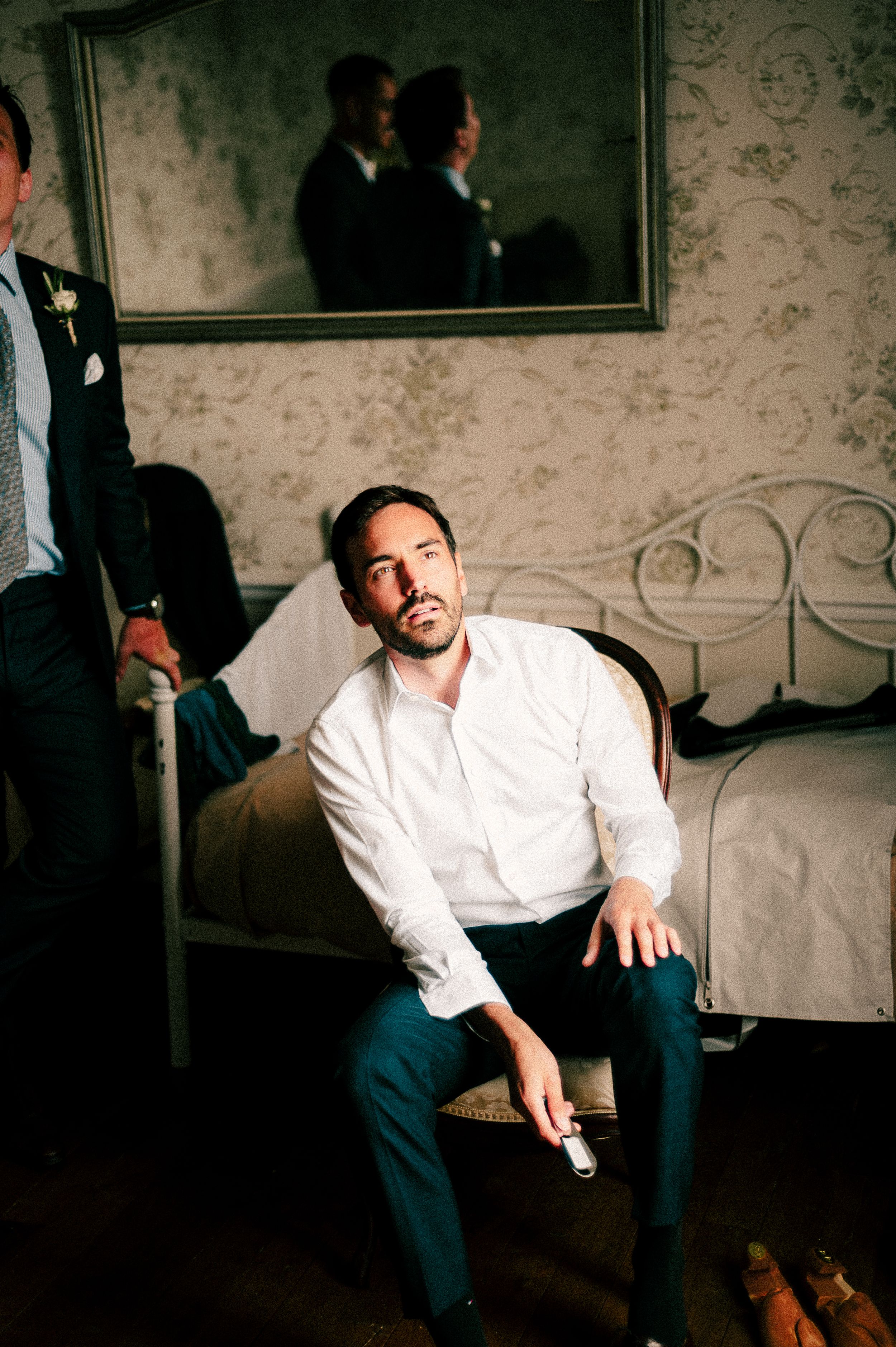 A person in formal attire sits on the edge of a bed in a vintage-style room with wallpaper and mirrors.