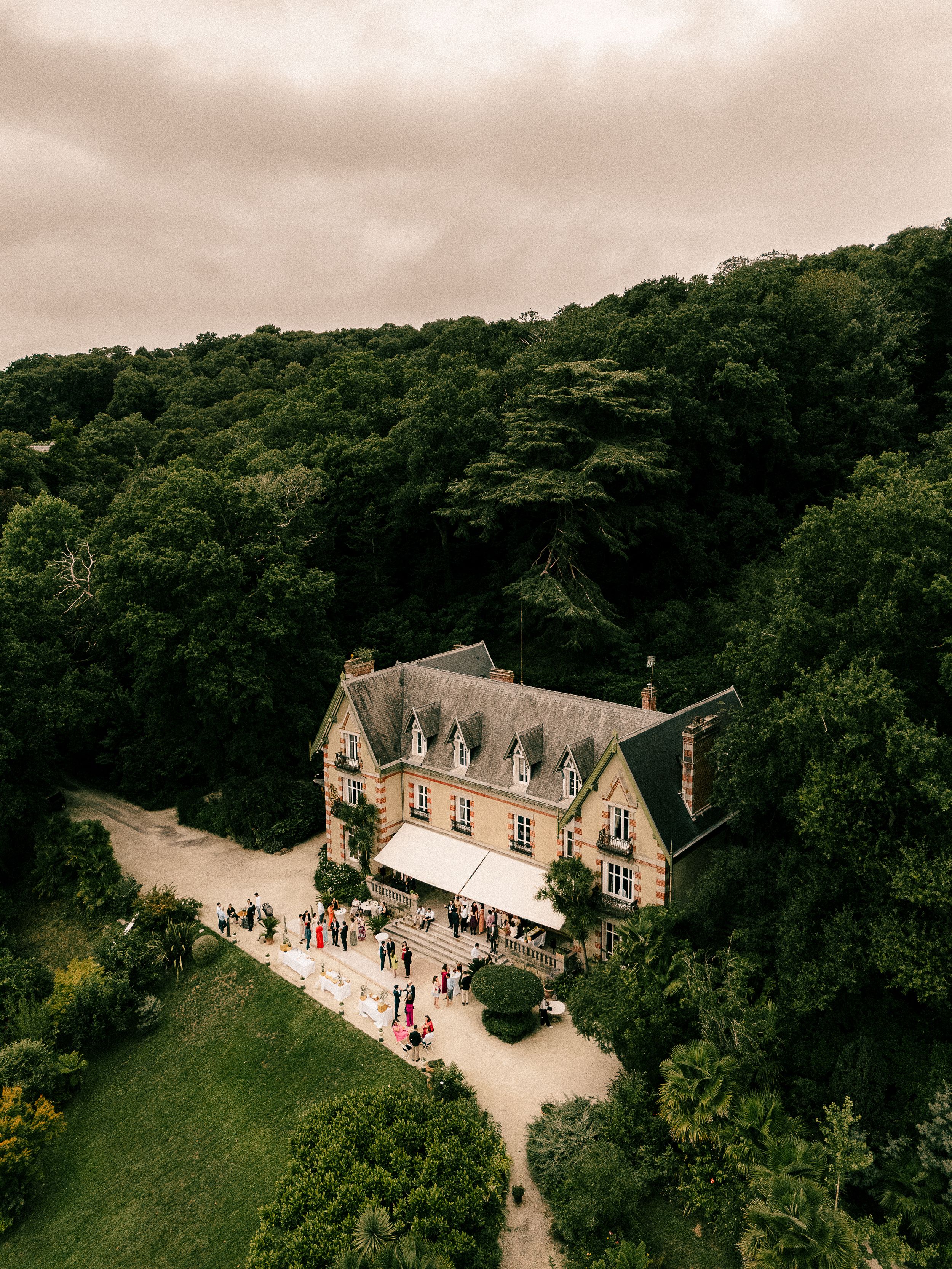 Aerial view of a grand stone manor house surrounded by lush forest with guests gathering outside.