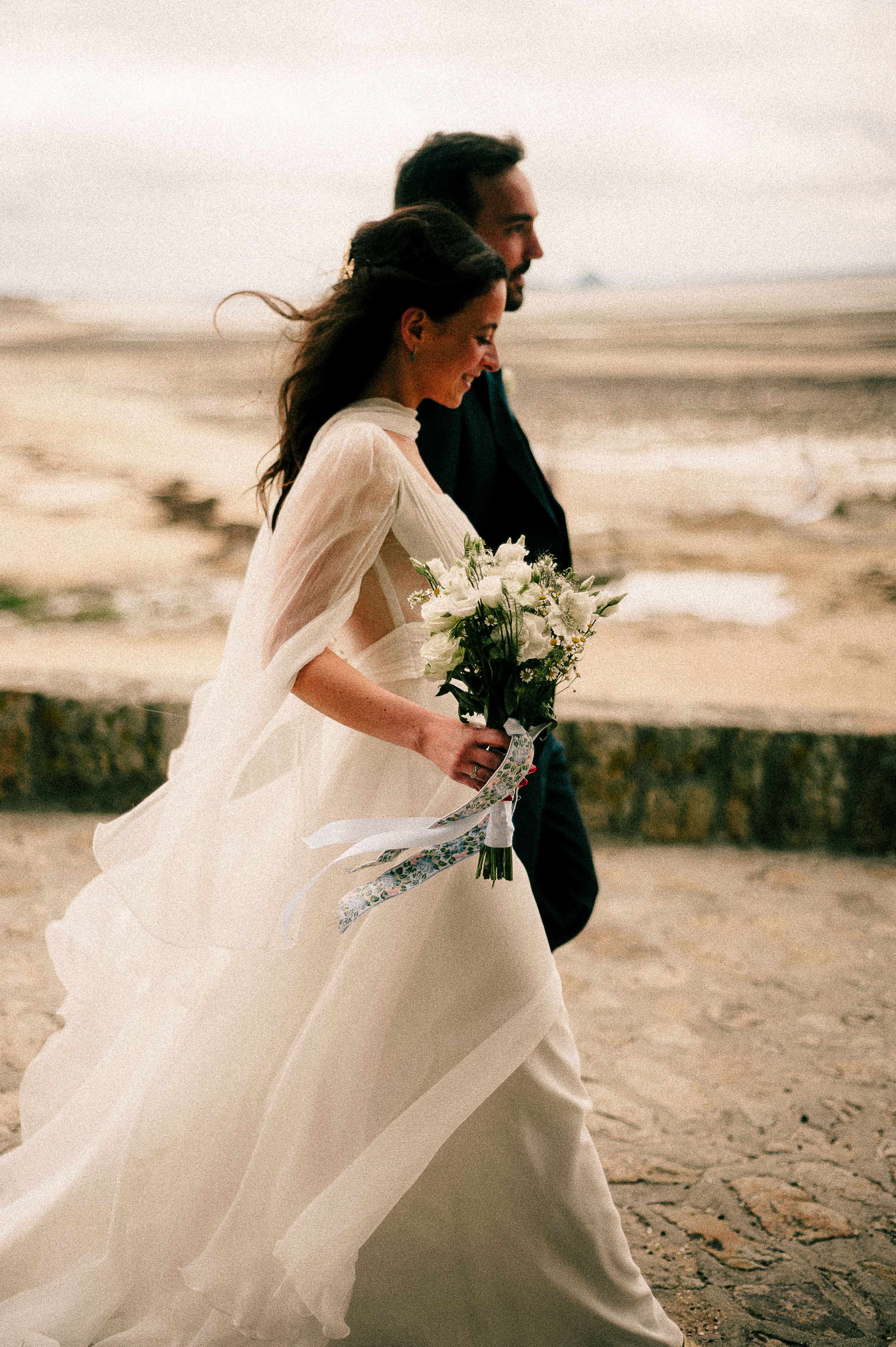 A romantic coastal wedding scene with a couple walking along a beach holding a white floral bouquet.