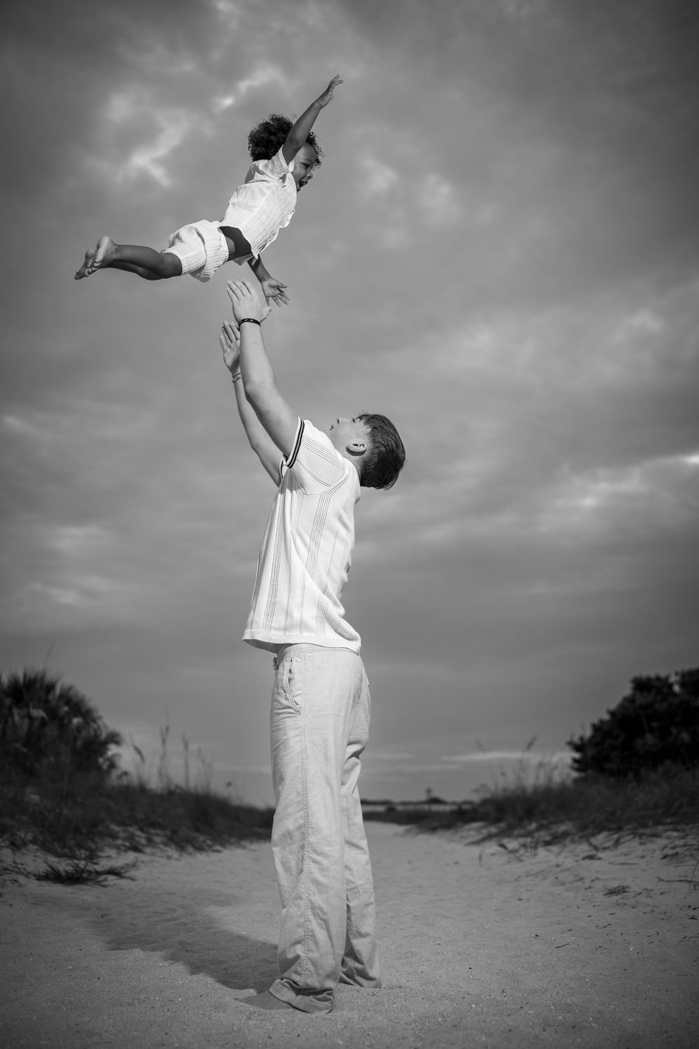 Black and white photo of parent lifting child high on sandy beach, artistic family photography in Tampa Bay, Florida.