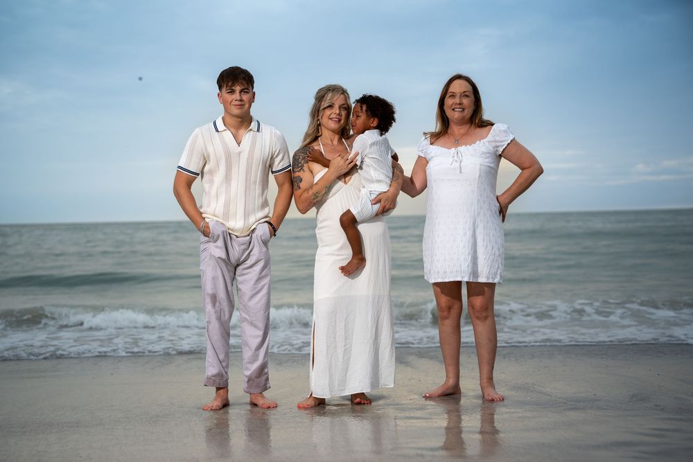 Family group portrait in light outfits standing on sandy beach with ocean waves, Tampa beach photography at golden hour.