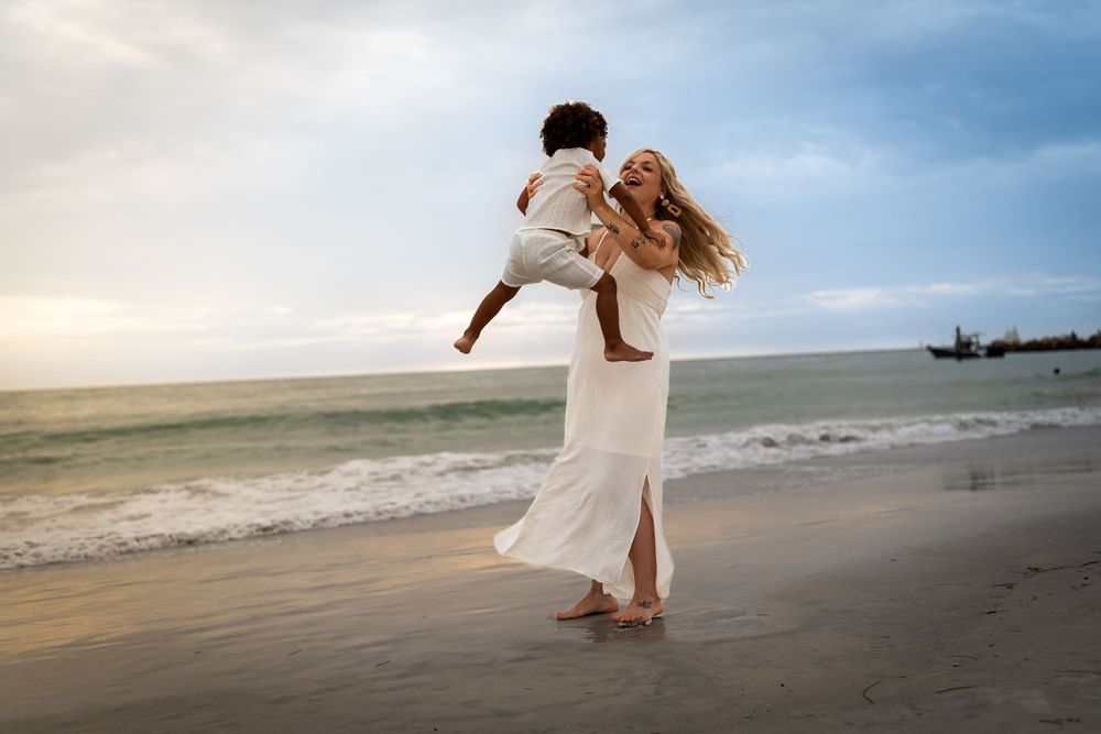 Mother in flowing white dress spinning joyfully with child on sunset beach, candid family photoshoot in Tampa, Florida.