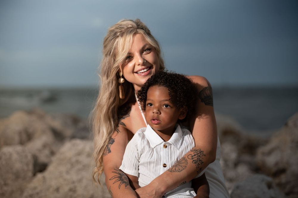 Mother embracing child on beach rocks, candid family photoshoot by the ocean in Tampa, Florida.