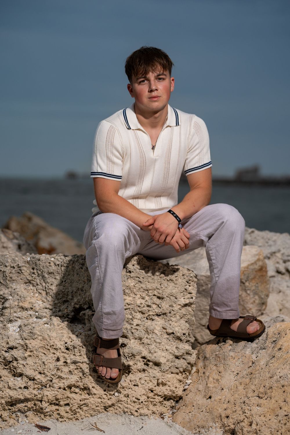 Man in white polo shirt and light pants sitting on rocky coastline at dusk, beach portrait photography in Tampa, Florida.