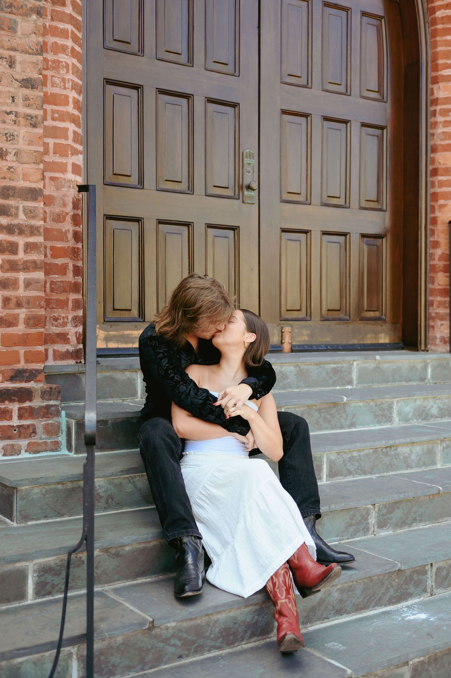A romantic couple sitting on stone steps in front of a large wooden door at a brick building.