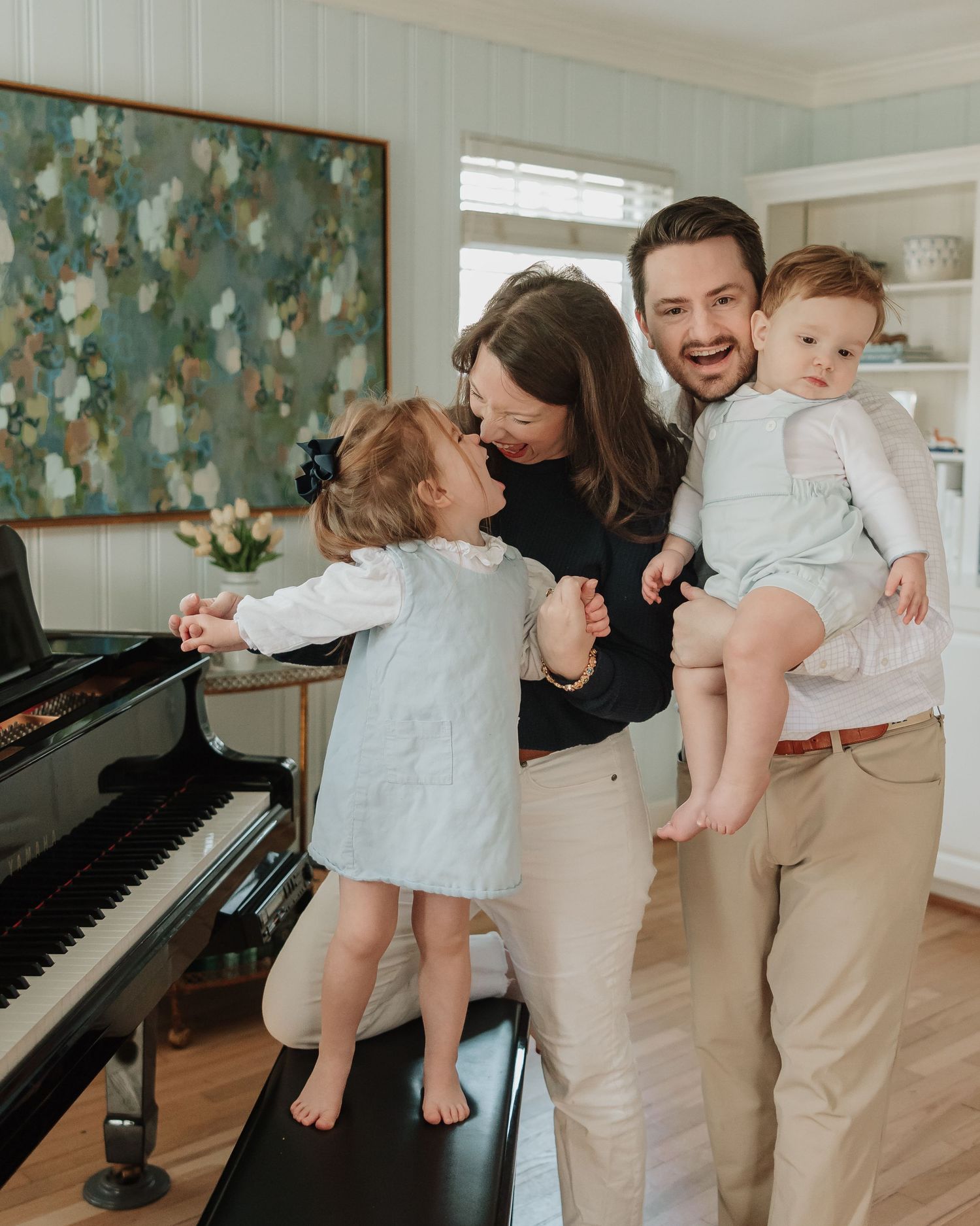 A mother and small child in light clothing share tender moments by a piano with blue patterned wallpaper in the background.