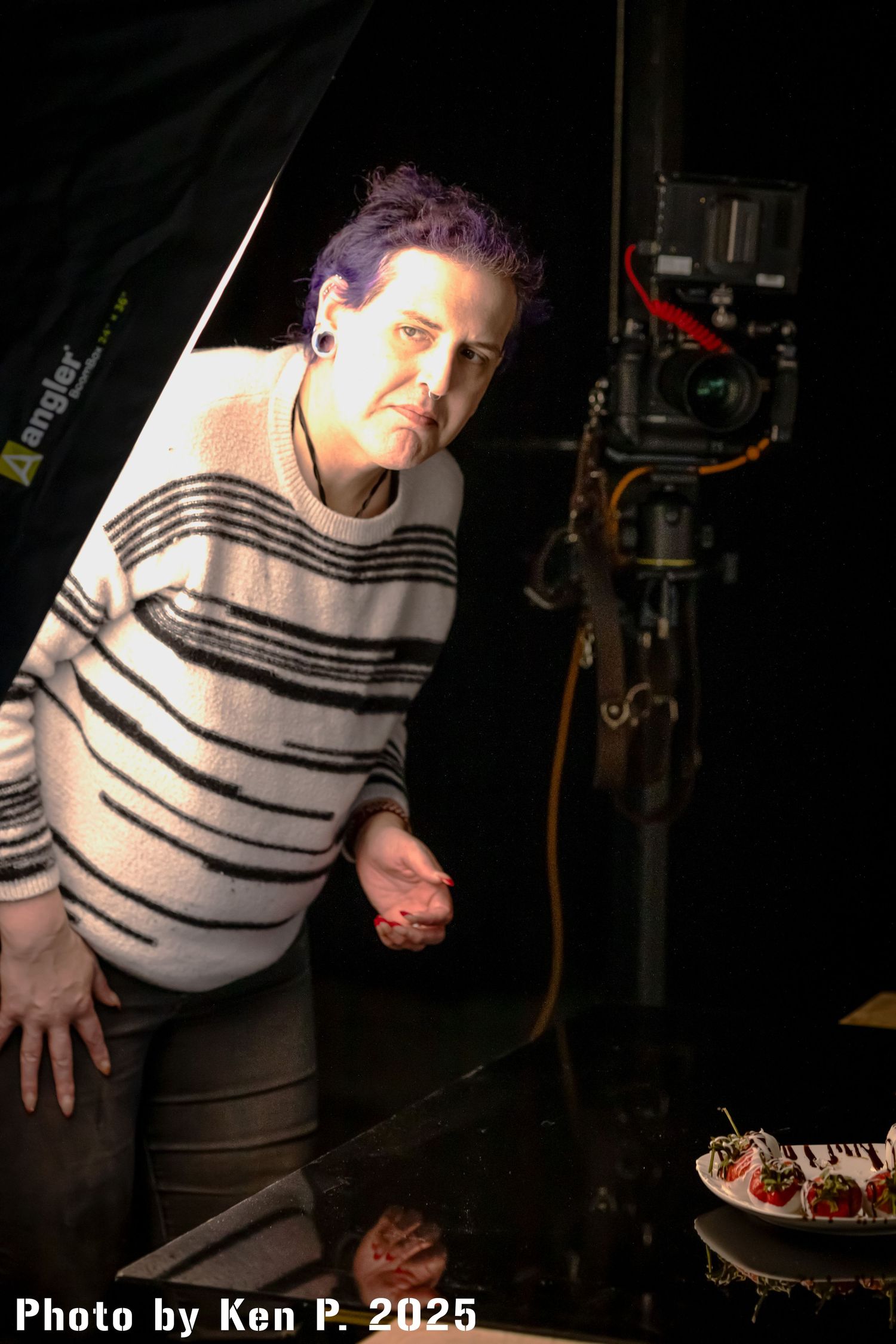 Catherine is standing in front of a studio light with a camera mounted on a tripod behind her with food decoration on a table and the lower