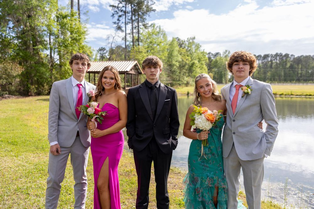 Five prom-dressed teenagers pose outdoors near a pond, wearing formal attire including pink and teal gowns and gray suits.