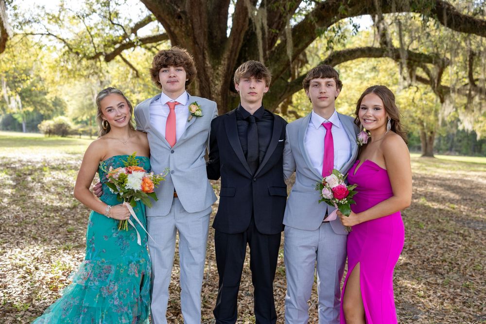 Five teens dressed in formal prom attire pose outdoors under a large oak tree, wearing suits and colorful gowns.