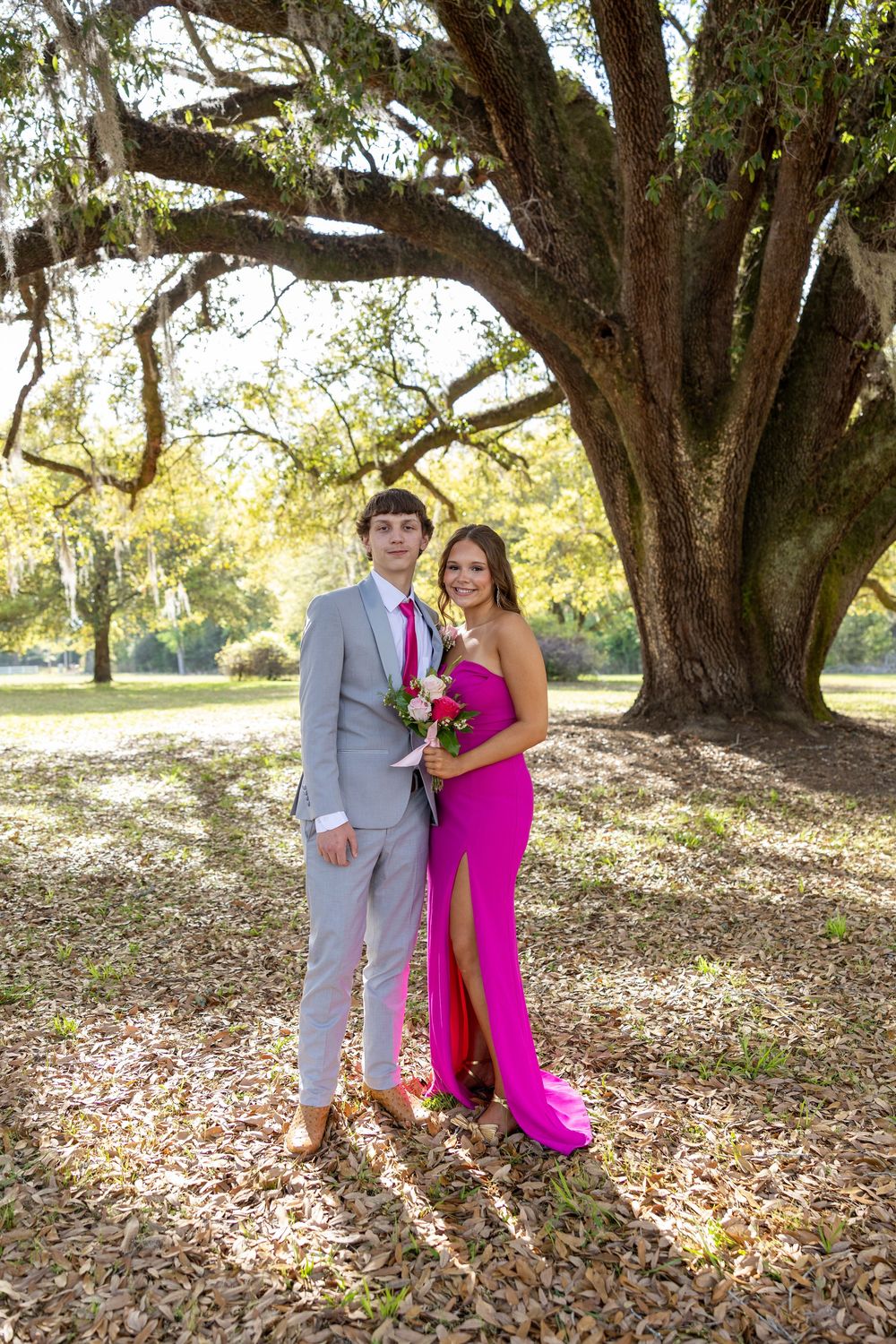 A couple in prom attire poses under a large oak tree, the girl in a pink gown and the boy in a gray suit.