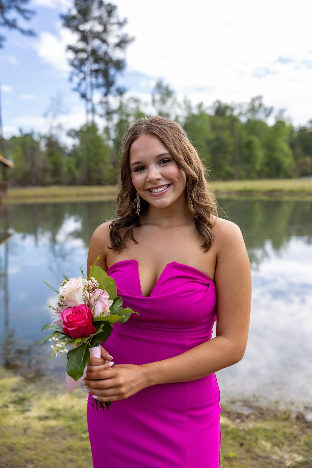 Smiling young woman in a magenta strapless gown holding a pink bouquet near a serene lake with trees.