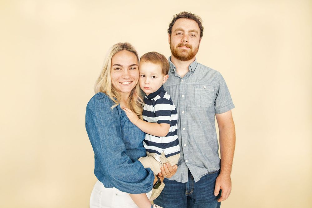 A smiling young family with a blonde mother, bearded father, and toddler son posing together on a beige background.
