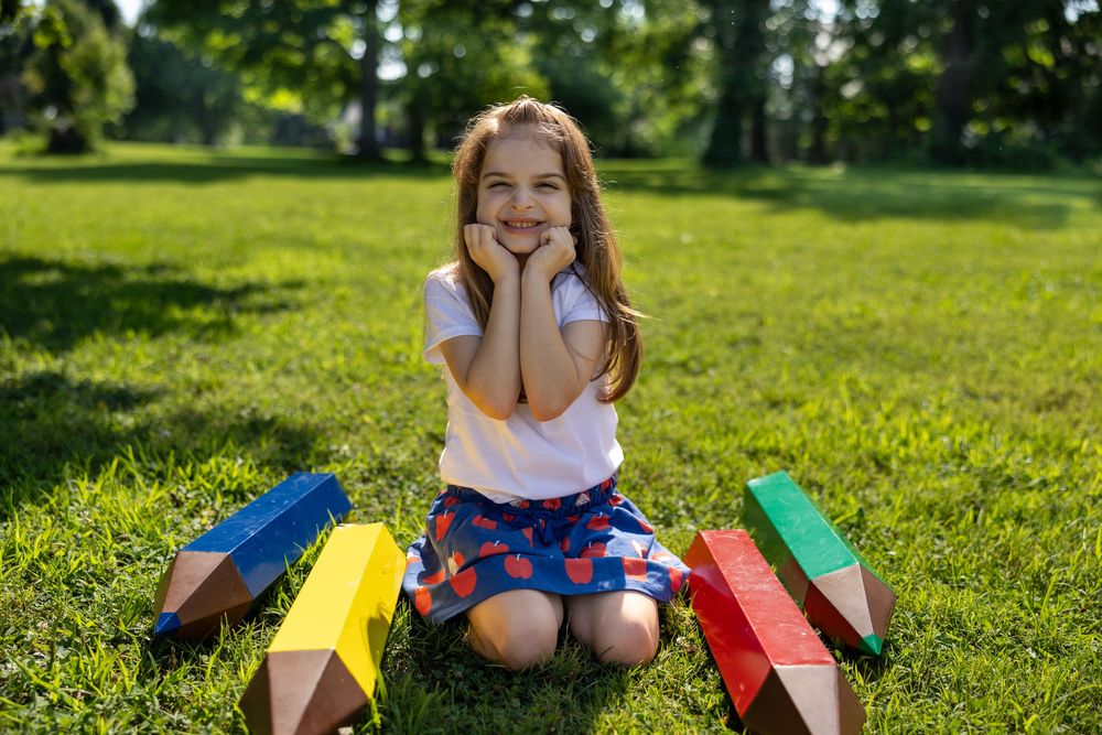 A happy little girl sits on green grass in a park surrounded by large colorful pencil props on a sunny day.