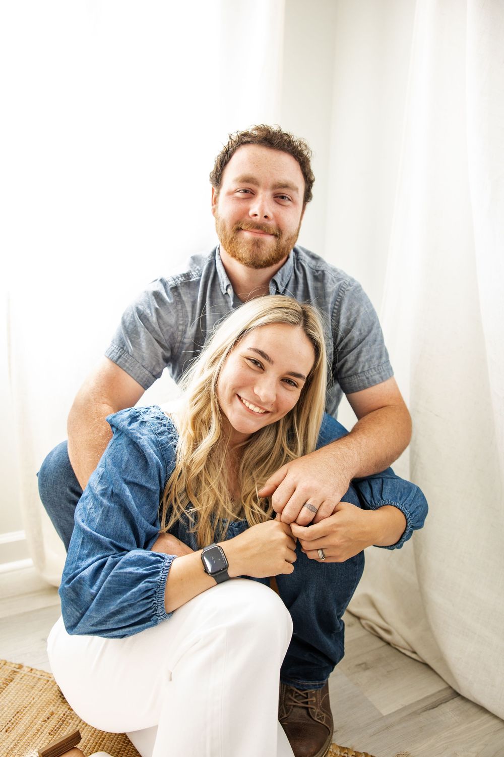 A smiling couple poses together indoors, the woman in a denim top and white pants, the man in a gray shirt and jeans.