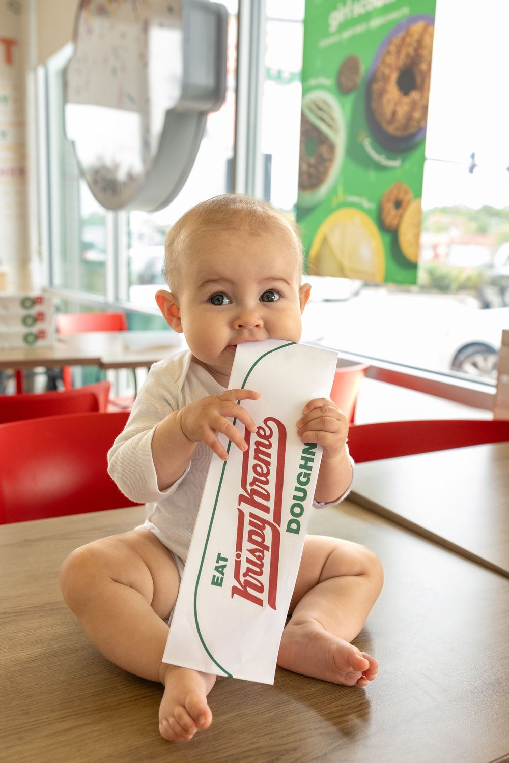 Adorable baby sitting on table chewing Krispy Kreme paper bag inside donut shop restaurant.