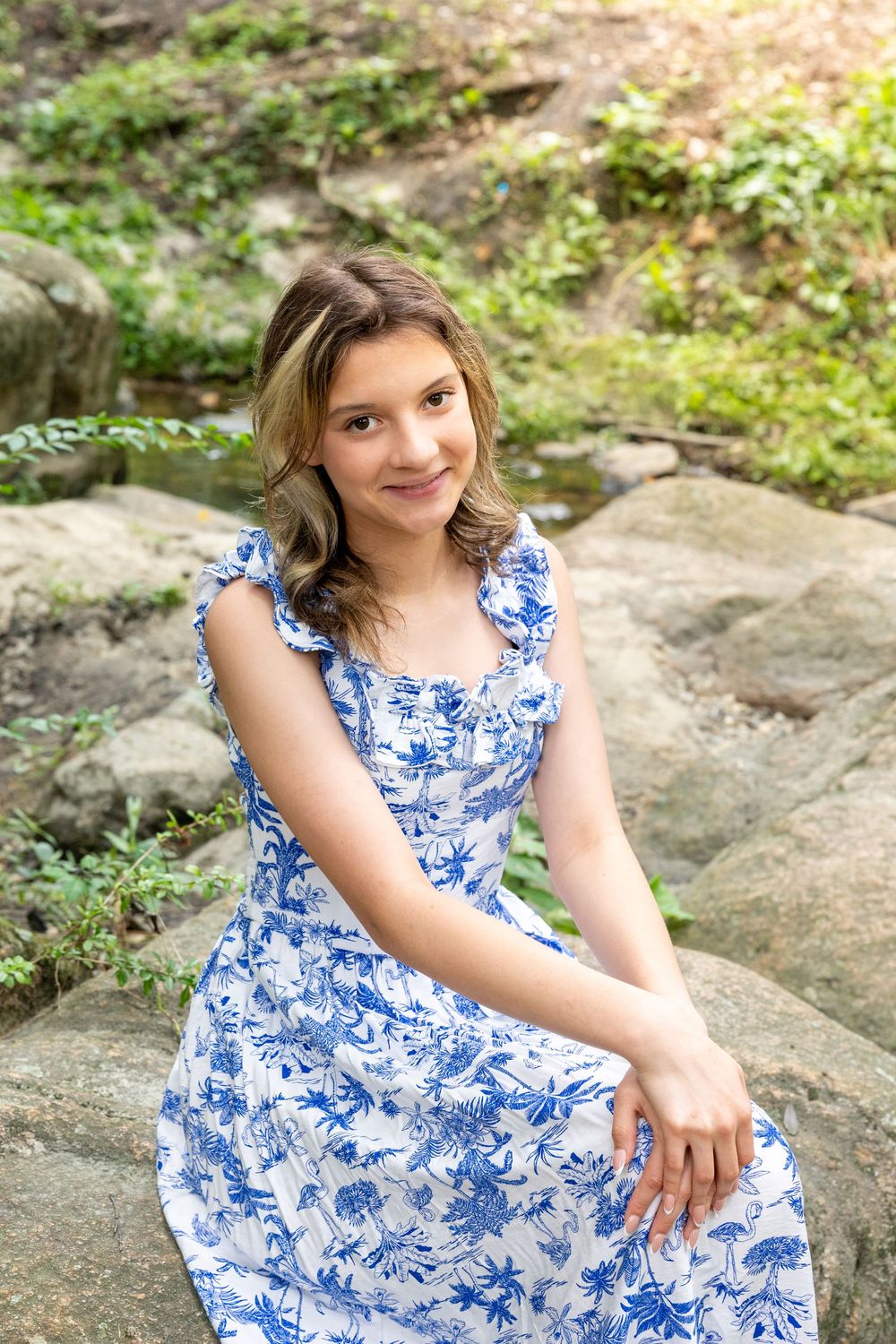 Young woman in blue floral dress sitting on rocks outdoors, smiling at camera with green foliage background.
