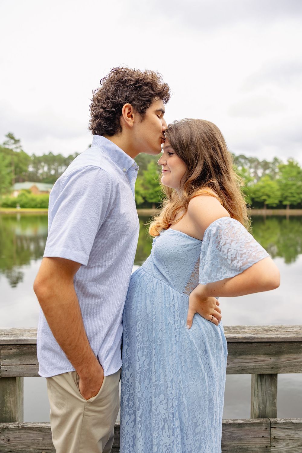 A couple shares a tender kiss on a wooden dock by a lake, the woman wearing a blue lace maternity dress.