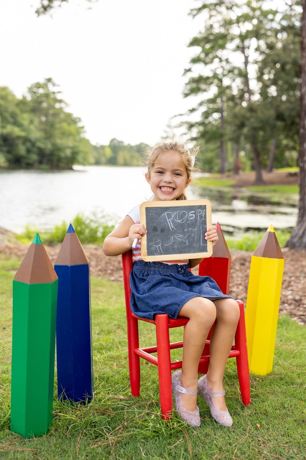 Young girl sitting on red chair holding chalkboard sign surrounded by giant colorful pencils outdoors near a lake.