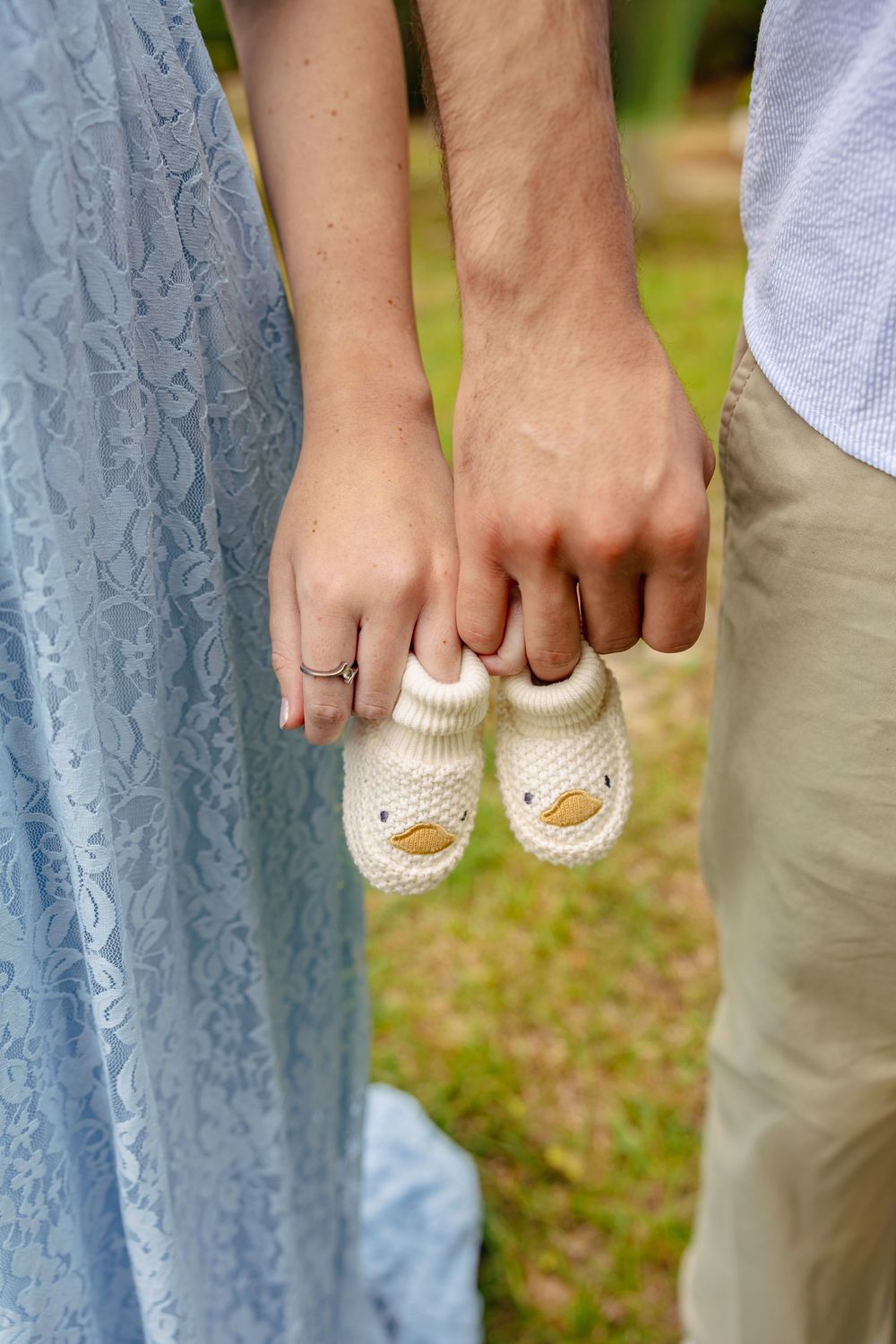 Couple holding tiny knitted duck baby booties together outdoors, pregnancy announcement photo with blue lace dress.