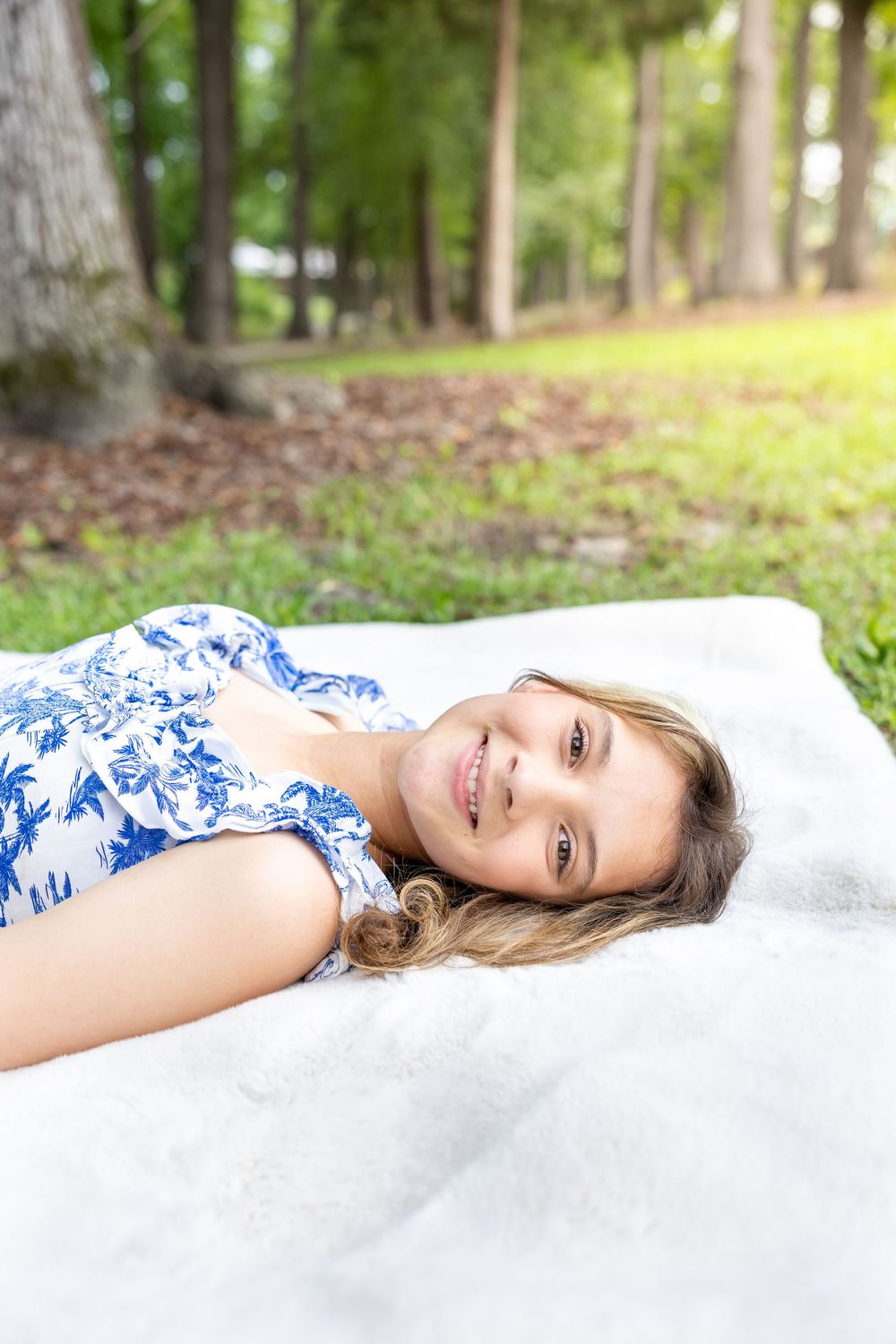 Young woman in blue floral dress lying on white blanket in park, smiling at camera.