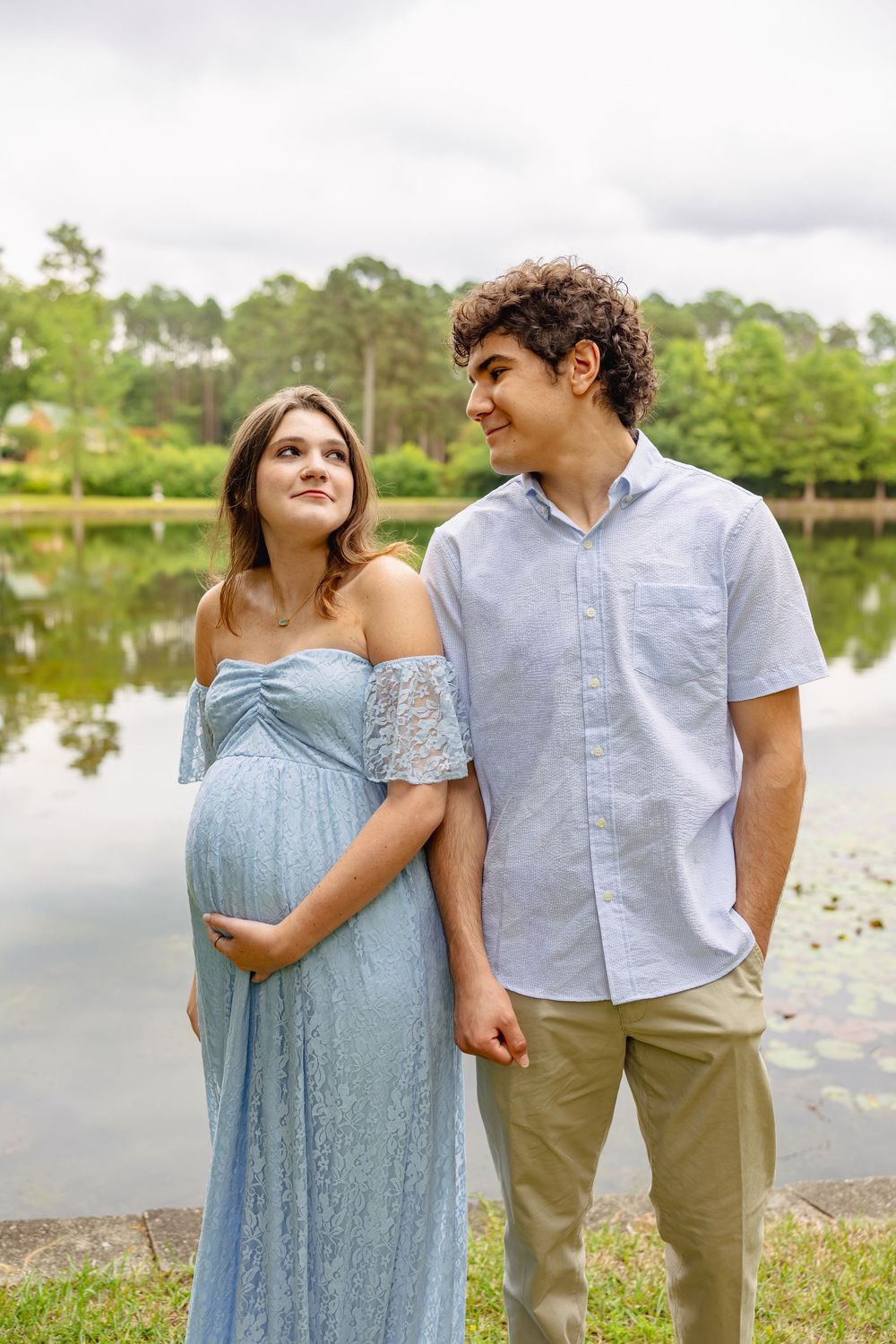 A pregnant woman in a blue lace dress stands with a man by a reflective lake, both smiling warmly.