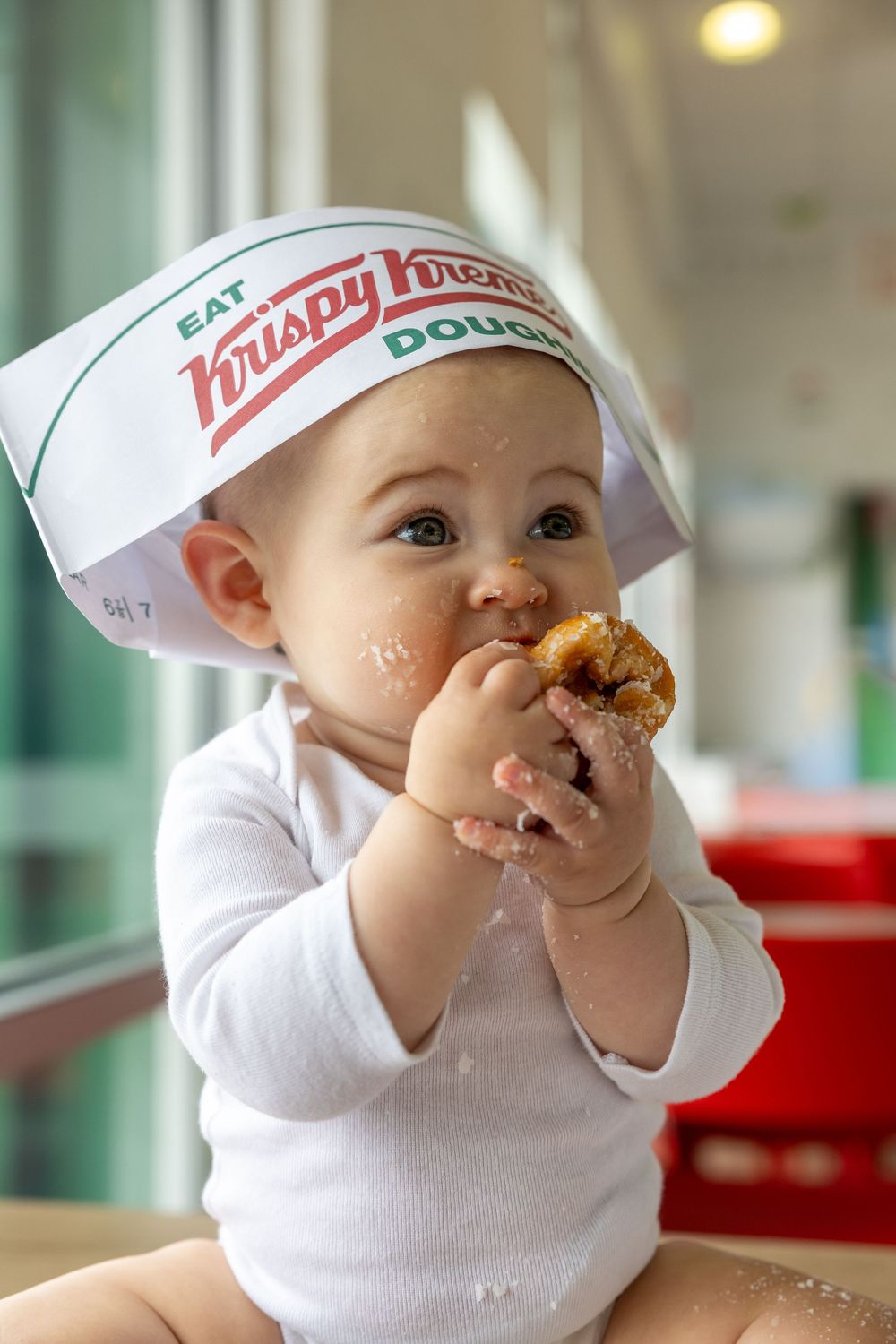 Adorable baby wearing a Krispy Kreme paper hat eating a glazed donut with frosting on their face.