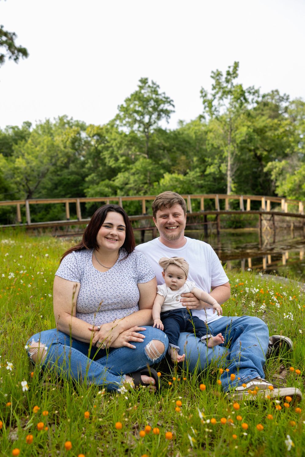 A happy family of three sits in a wildflower meadow, with parents holding their baby girl near a wooden bridge.