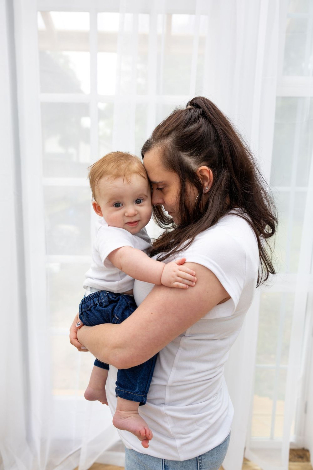 A loving mother holds her smiling red-haired baby, nuzzling close near a bright white curtained window.