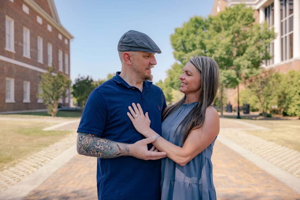 A couple shares a romantic moment outdoors, the man with tattoos wearing a blue polo and flat cap, the woman in a gray dress.