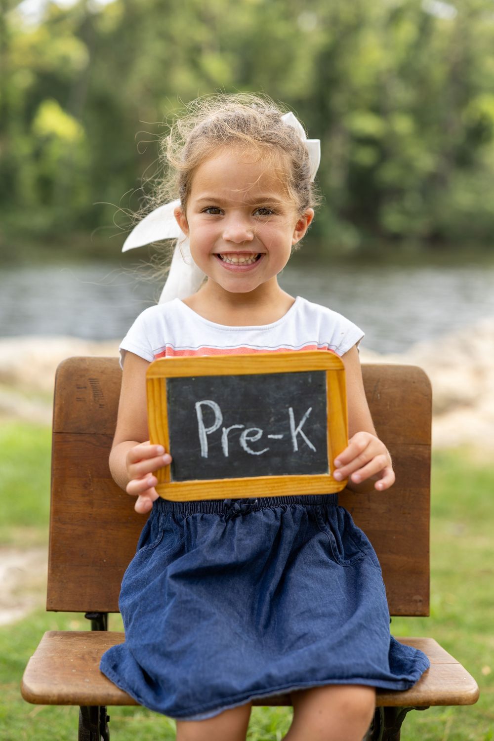 Smiling young girl sitting on a bench outdoors, holding a chalkboard sign that reads Pre-K.