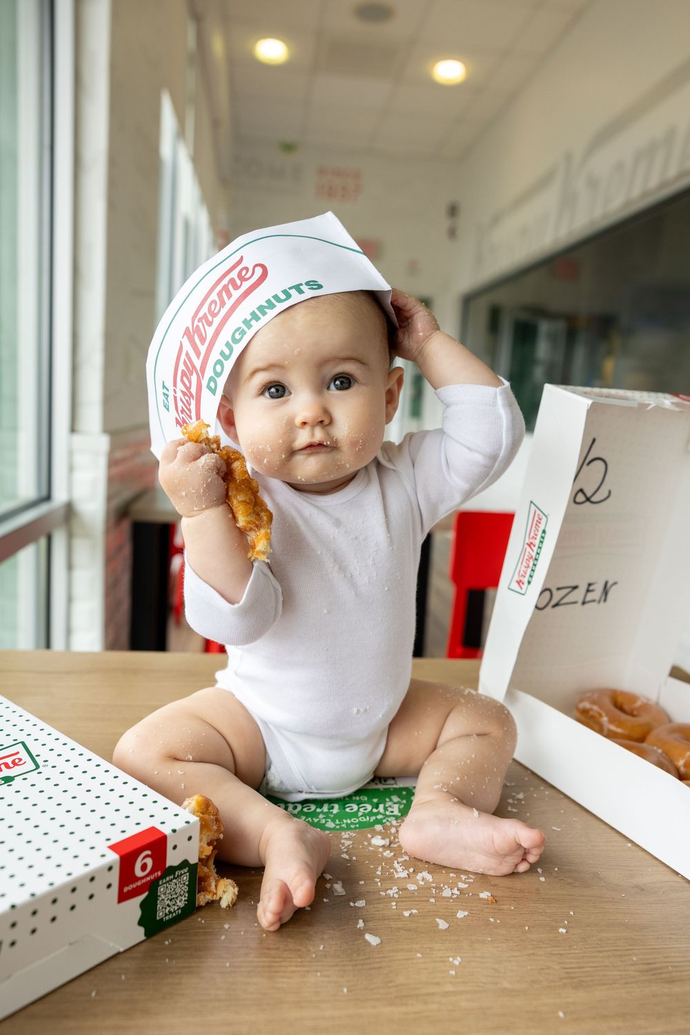 Adorable baby wearing Krispy Kreme paper hat eating a donut while sitting on table with open donut boxes.