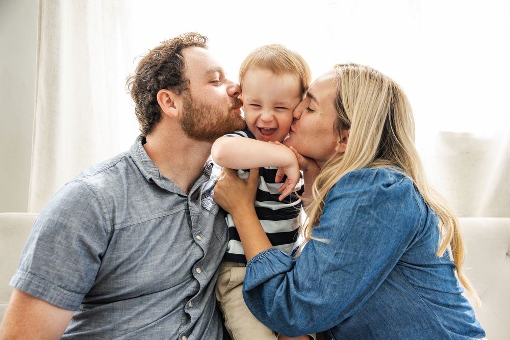 Happy parents kissing their laughing toddler on the cheeks while sitting together on a couch at home.