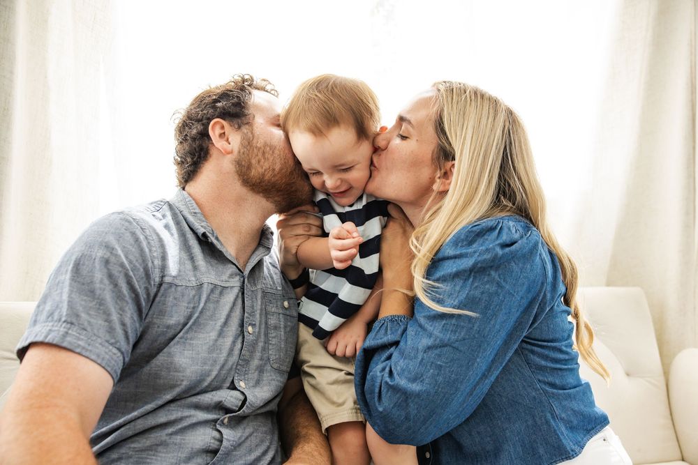Happy parents kissing their smiling baby boy on the cheeks while sitting together on a white couch.