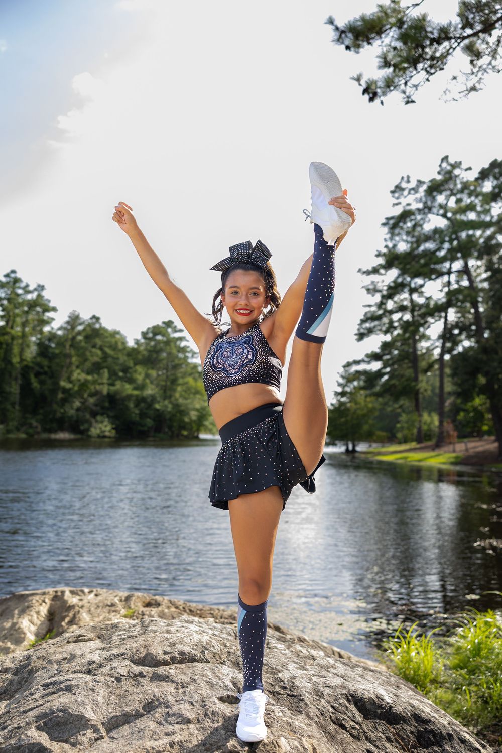 Cheerleader in navy uniform performs high kick on rocks by a lake surrounded by trees.