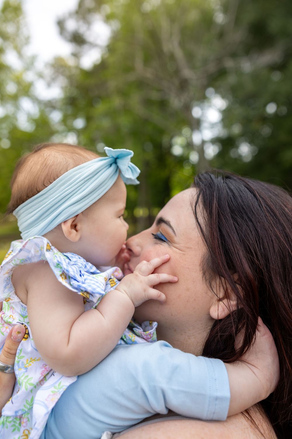 A mother holds her baby girl wearing a blue bow headband outdoors, as the infant playfully touches her face.