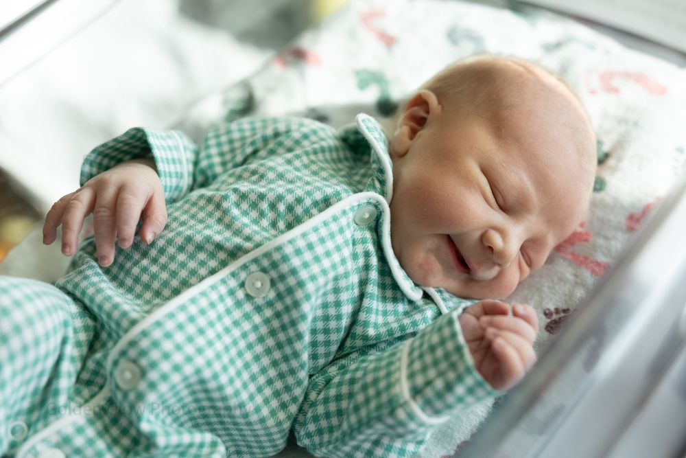 Newborn baby sleeping peacefully in green gingham pajamas in a hospital bassinet.