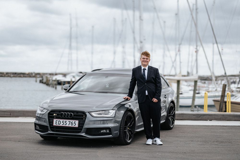 A sleek grey Audi S4 parked at a marina with sailboat masts visible in the background.
