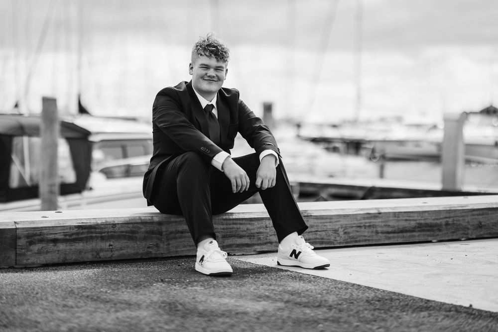 Black and white portrait of someone in a suit sitting casually on a wooden bench at a marina with boats in background.