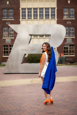 Boise State University graduate of nursing holding blue gown over shoulder, looking back at camera in front of the B, wearing orange heels
