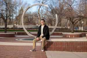 Male Boise State University graduate in street clothes sitting near the circular sculpture on campus