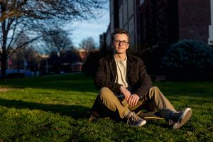 Male Boise State University graduate wearing street clothes sitting on skateboard in grass