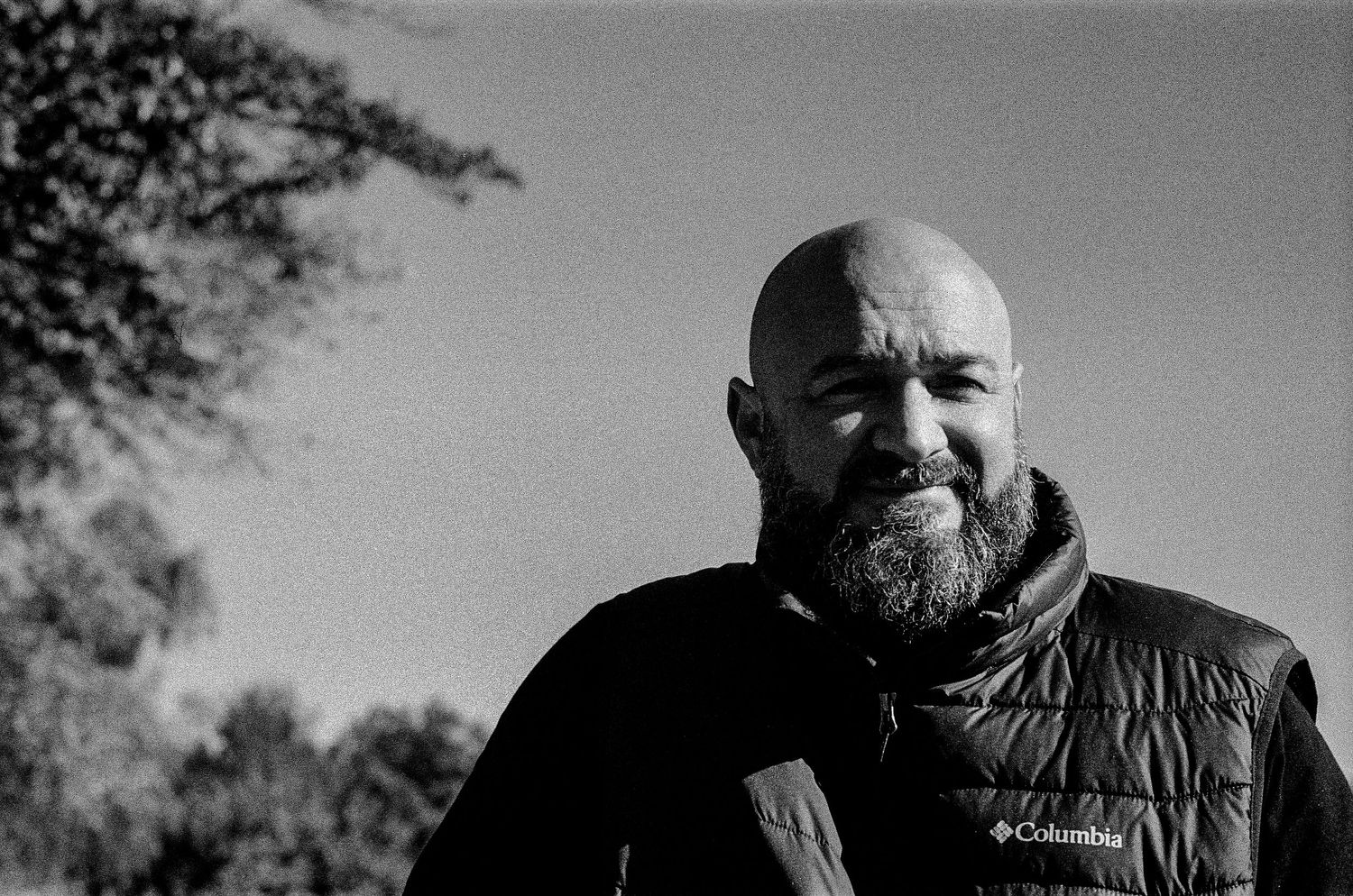 Black and white portrait of person wearing a Columbia vest outdoors with trees in background.