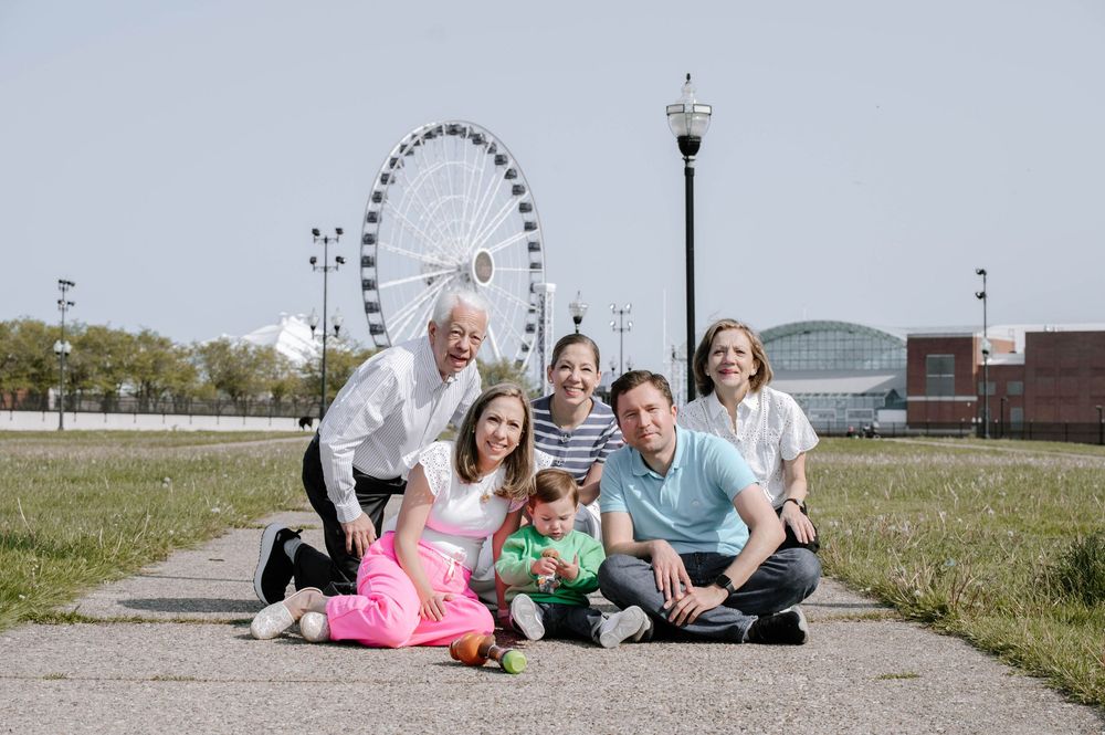 Happy family group photo outdoors near a Ferris wheel on a sunny day, smiling and posing together on a path.